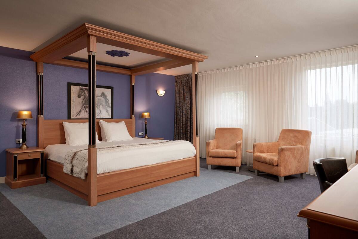 Ein Bett mit Baldachin und Holzrahmen, zwei Nachttische jeweils mit Lampe, ein großes Fenster mit durchsichtigen Vorhängen sowie zwei Sessel.