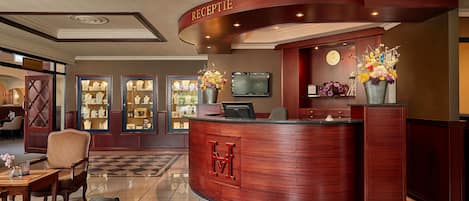 A hotel reception area with a curved wooden reception desk, a clock, a television, and a vase of flowers.