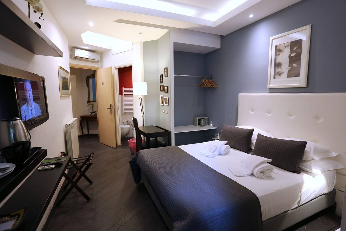 A hotel room with a large bed, a desk with a computer, a TV, and a kitchenette.