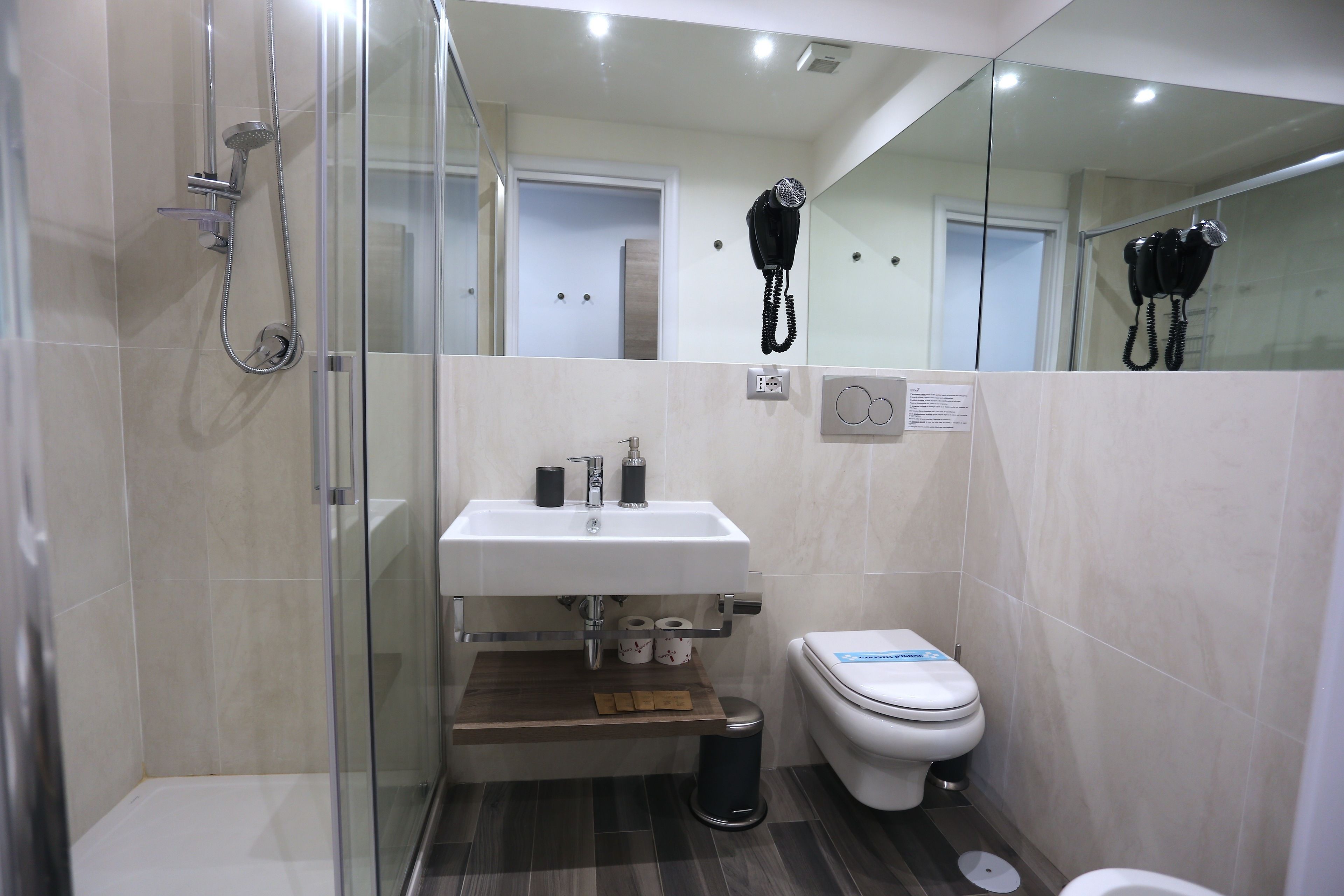Baño pequeño con ducha de vidrio, lavamanos blanco con espejo arriba y un inodoro con un adhesivo azul.