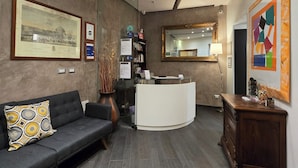 A reception area with a reception desk, a sofa, a vase with branches, a mirror, and framed artwork on the walls. - B&B Home 77 (Rome)