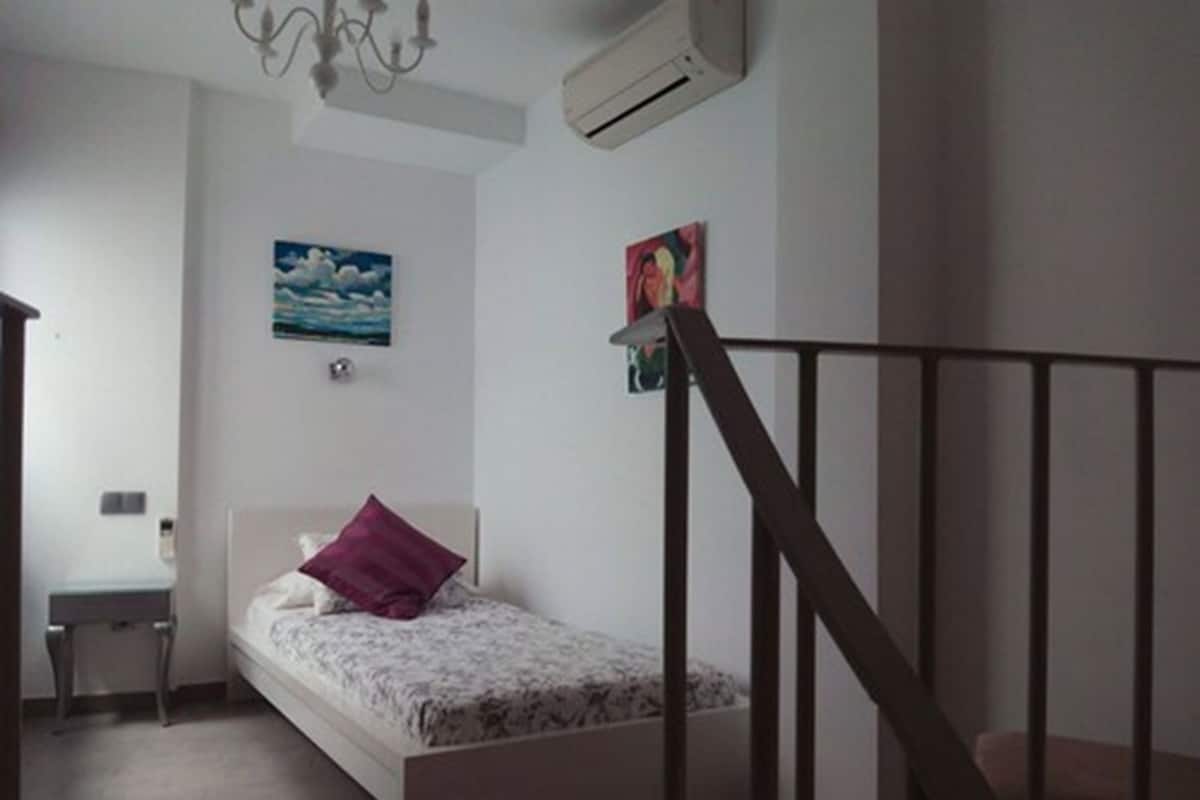 A bedroom with a bed, a desk, and a chair. There is a painting on the wall and an air conditioning unit above the bed.