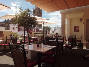 An outdoor cafe with tables and chairs, a tree, and a view of a historic building in the background. - Hotel Palacio Alcazar (Seville)