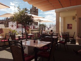 Un café al aire libre con mesas y sillas, un árbol y una vista de un edificio histórico al fondo.