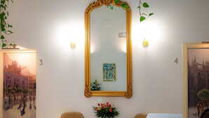 A hotel room with a large ornate mirror, two chairs, a small table with a vase of flowers, and two paintings on the walls. - Hotel Palacio Alcazar (Seville)