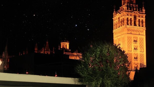 A night view of a terrace with wicker chairs and a table, overlooking a historic tower illuminated against the night sky. - Hotel Palacio Alcazar (Seville)