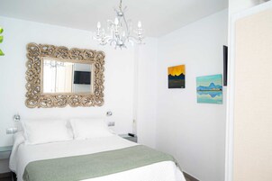A hotel room with a large ornate mirror, a bed with white linens, a green throw blanket, a chandelier, and two paintings on the wall.