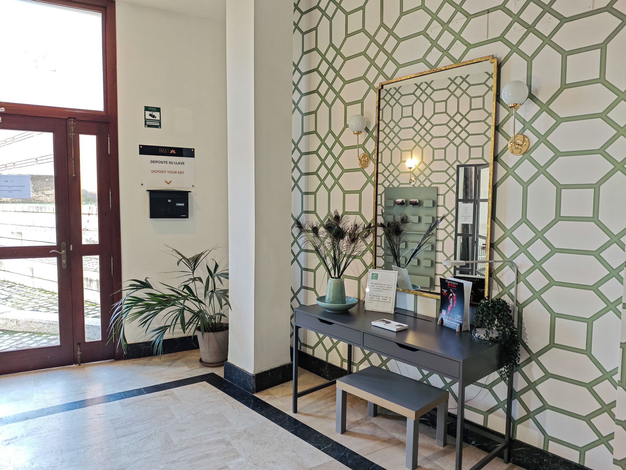 A reception area with a desk, bench, and decorative plants. There is a door with a sign above it, and a wall with a geometric pattern.