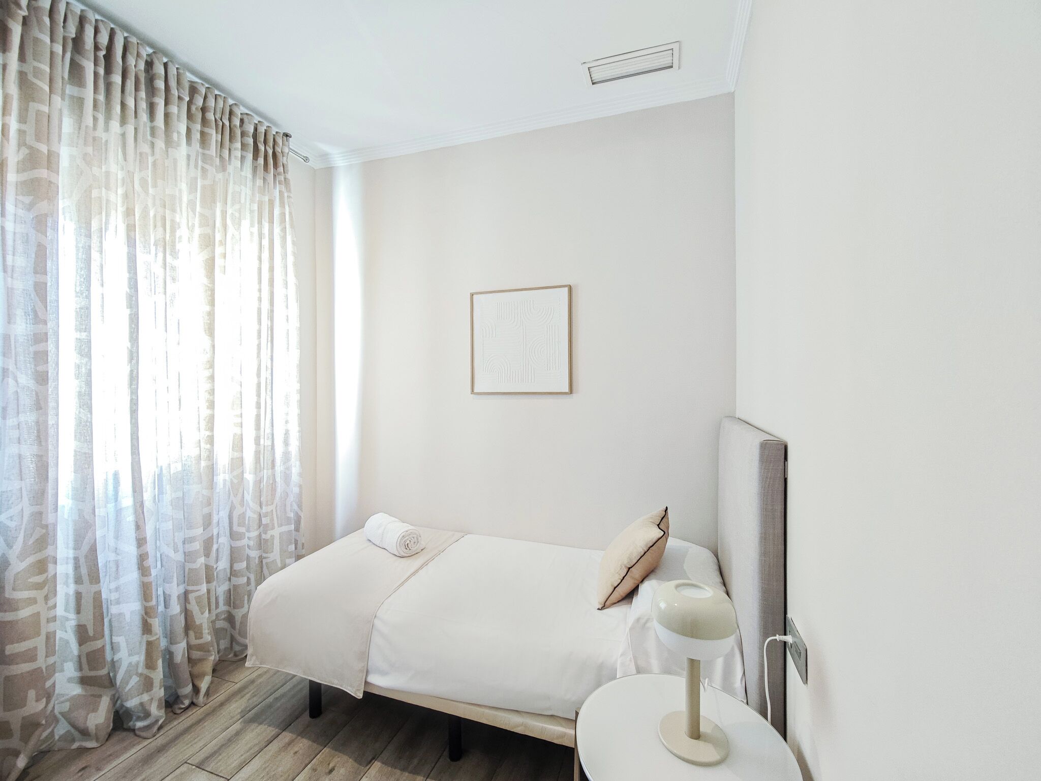 Una habitación de hotel con cama, mesita de noche, un cuadro en la pared y una ventana con cortinas.