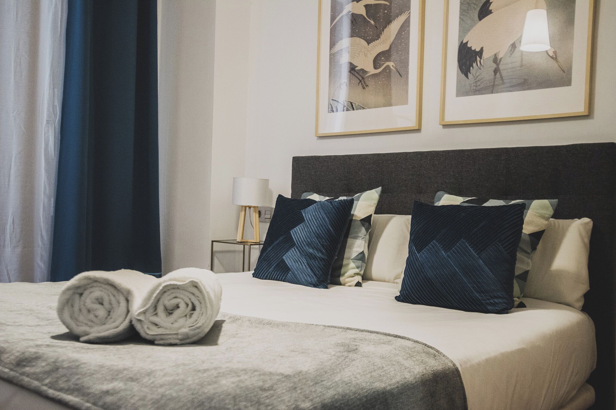 Una cama bien tendida con toallas enrolladas, una lámpara y dos cuadros en la pared.