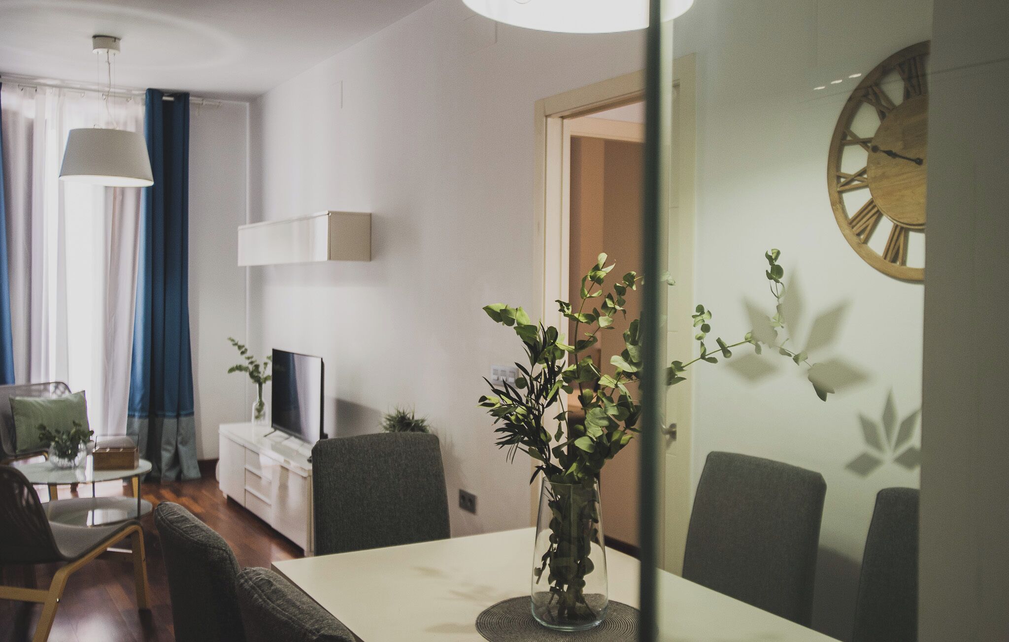 A modern living room with a dining table, chairs, a TV, and a clock on the wall.