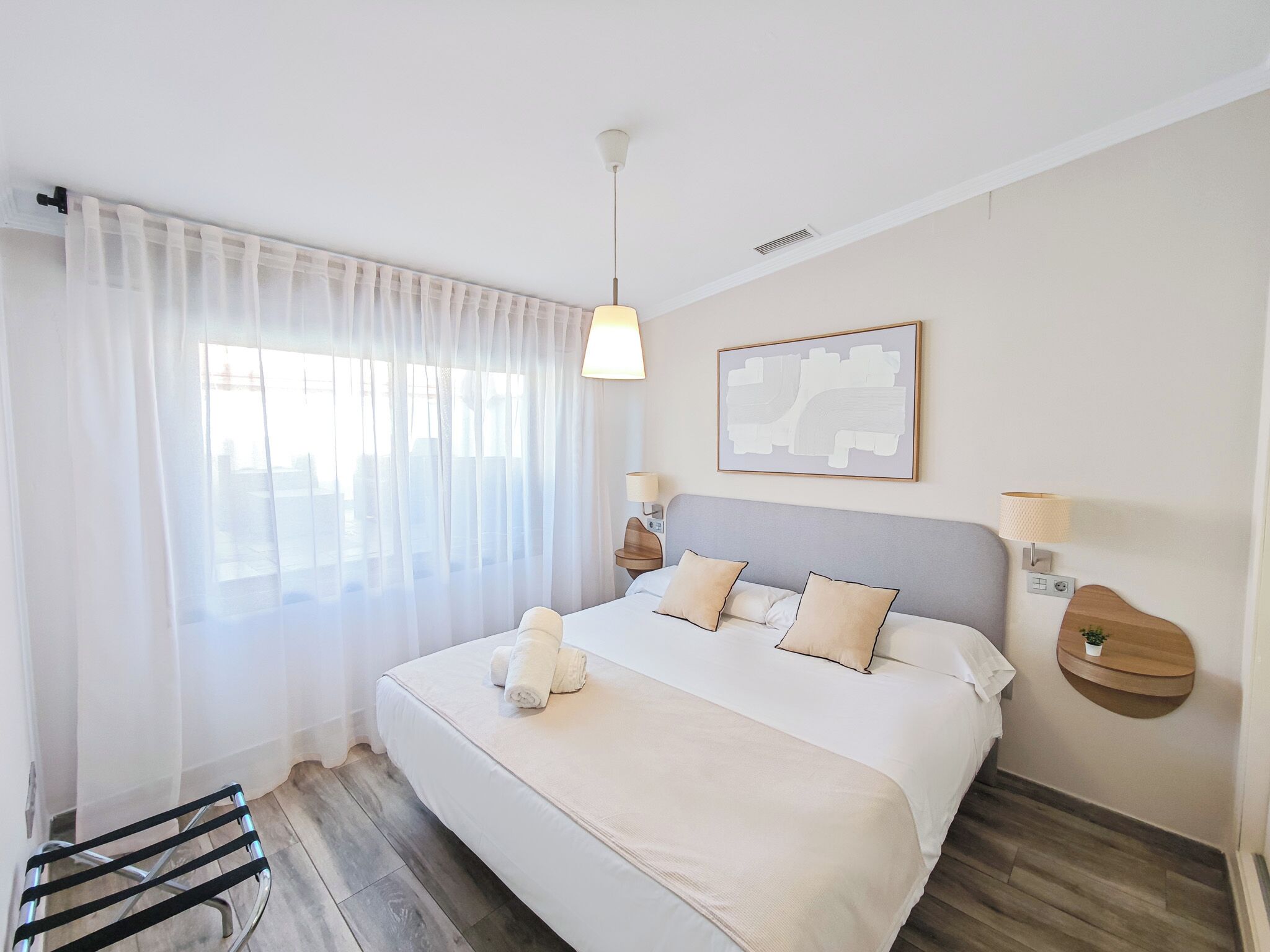 A hotel room with a large bed, a window with sheer curtains, a bedside table with a lamp, and a wall-mounted artwork.