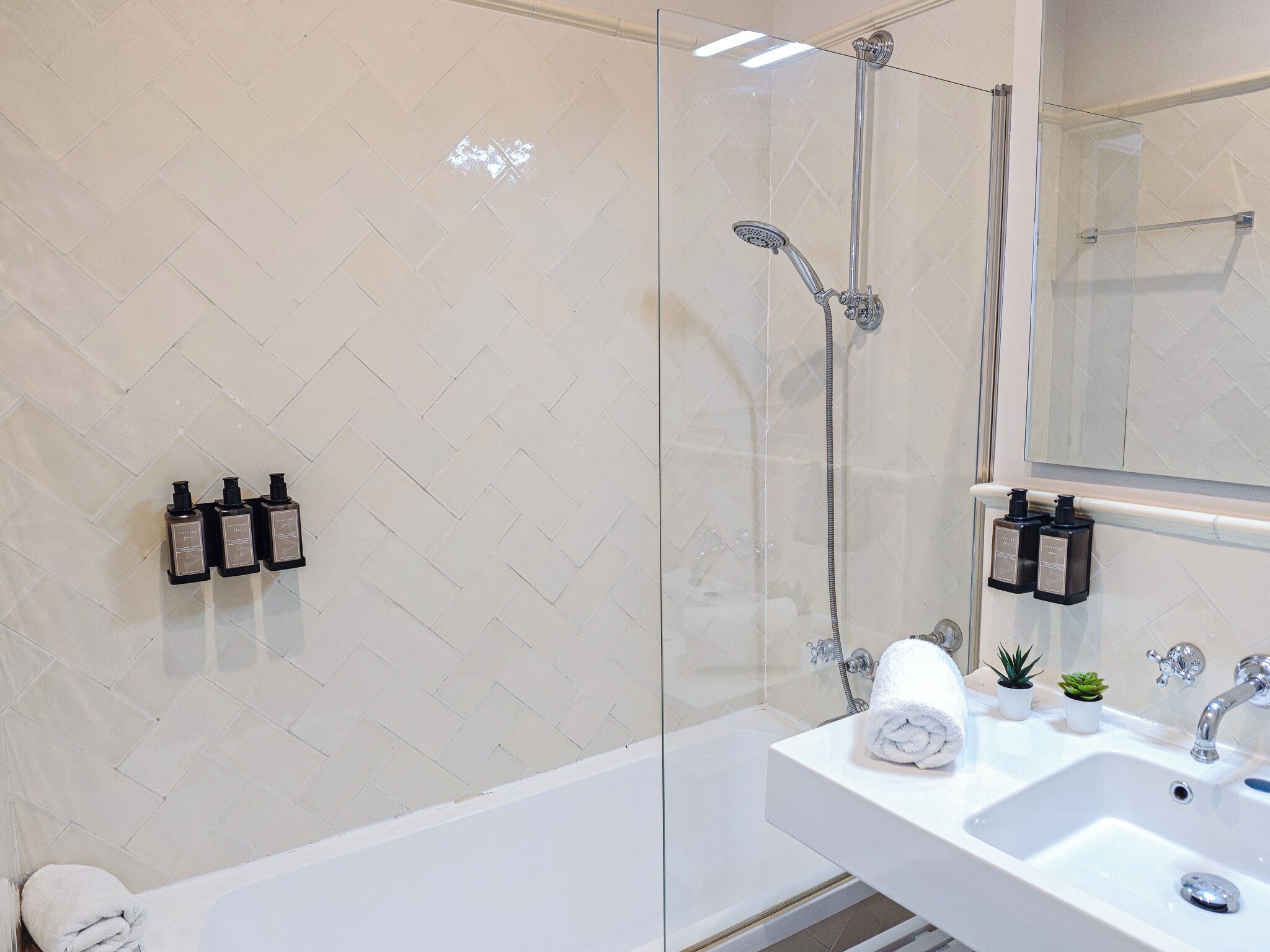 Un baño moderno con cabina de ducha de vidrio, una bañera blanca y un lavamanos con una toalla y dos pequeñas macetas.