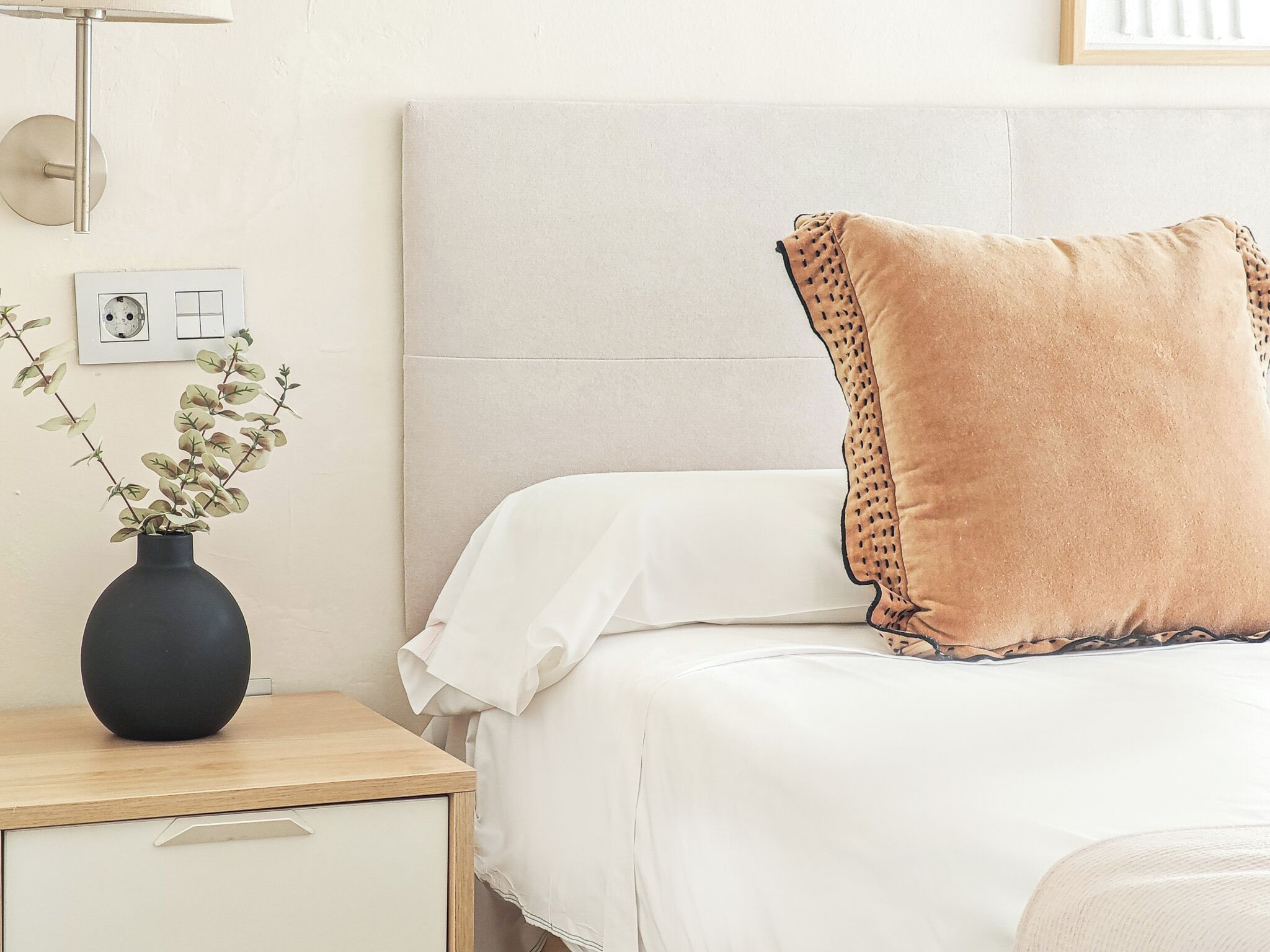 Una cama bien hecha con una colcha blanca y un almohadón marrón con detalle decorativo. Un jarrón negro con follaje verde se encuentra sobre una mesita de noche de madera junto a la cama.