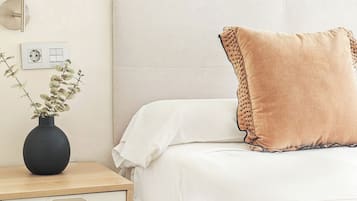 Una cama bien hecha con una colcha blanca y un almohadón marrón con detalle decorativo. Un jarrón negro con follaje verde se encuentra sobre una mesita de noche de madera junto a la cama.