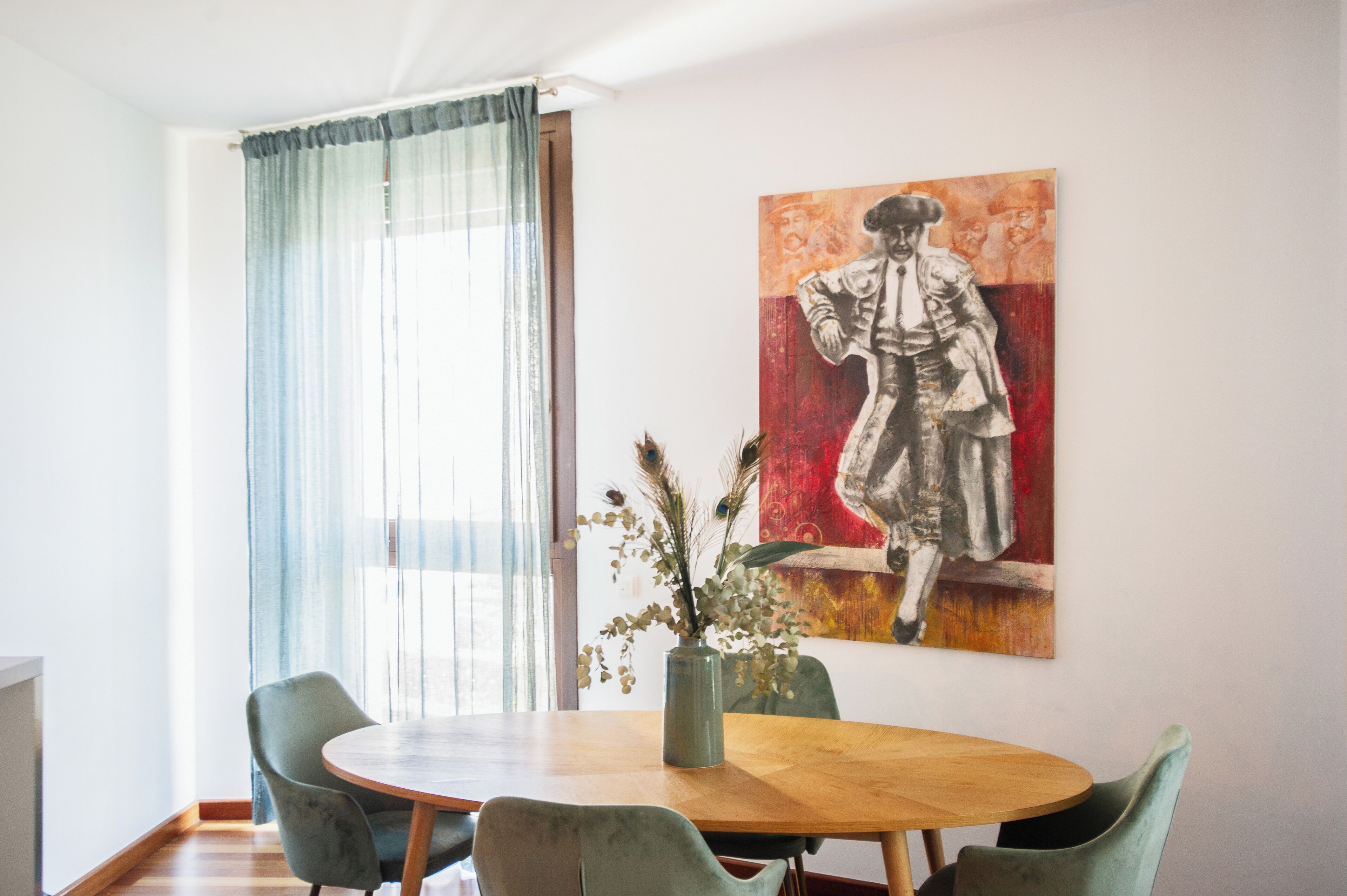 A dining area with a wooden table, four chairs, a vase with flowers, and a large painting on the wall.