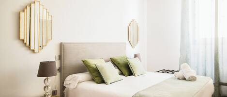 Una cama bien hecha con cabecera blanca, dos almohadas verdes y una toalla blanca al lado. Hay una mesita de noche con una lámpara junto a la cama.