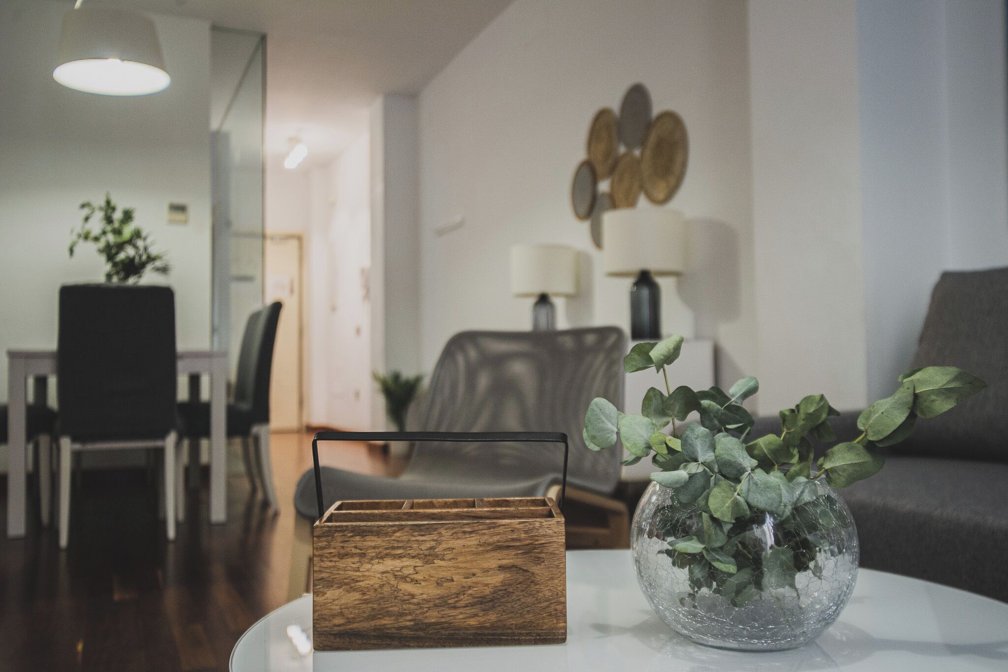 A modern living room with a wooden side table, a glass vase with green foliage, a grey sofa, and a dining area in the background.