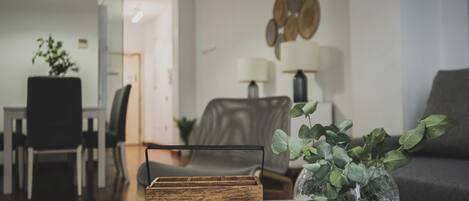 A modern living room with a wooden side table, a glass vase with green foliage, a grey sofa, and a dining area in the background.