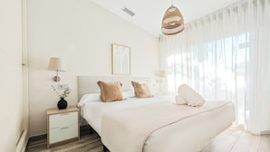 A modern bedroom with a large bed, bedside lamps, a bedside table with a lamp, a hanging light fixture, and a window with sheer curtains. - AZZ Sevilla Torre de la Plata (Seville)