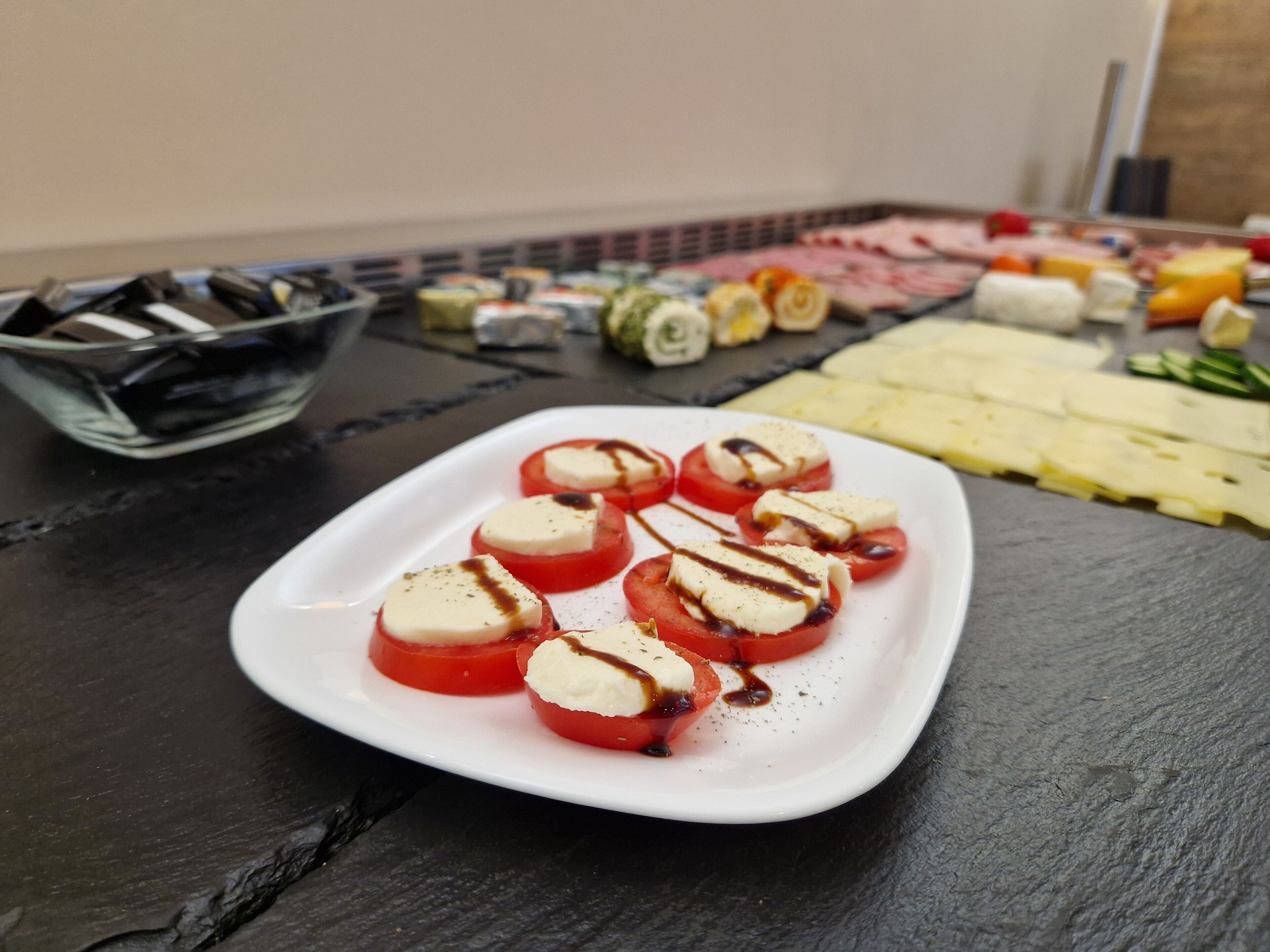 Un piatto bianco con fette di pomodoro, formaggio e salsa, una ciotola di sushi e diversi altri cibi su una superficie scura.