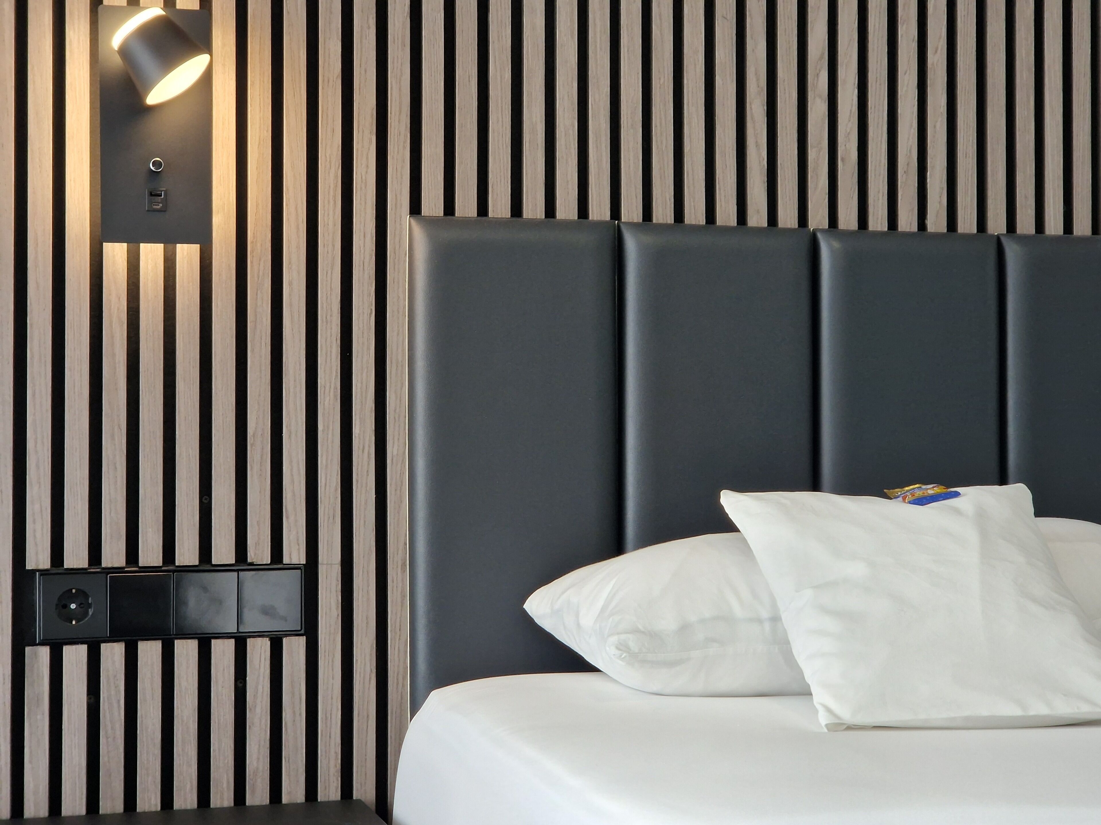 Ein modernes Hotelzimmer mit einer Wandpaneele in Holzoptik mit Streifen, einem dunklen Kopfteil und einem Bett mit weißer Bettwäsche und Kissen.