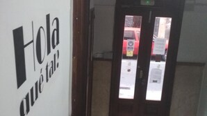 A hallway with a sign reading "Hola qué tal?" and a door with a view of a red car outside. - Pensión Sea of Clouds (Las Palmas de Gran Canaria)