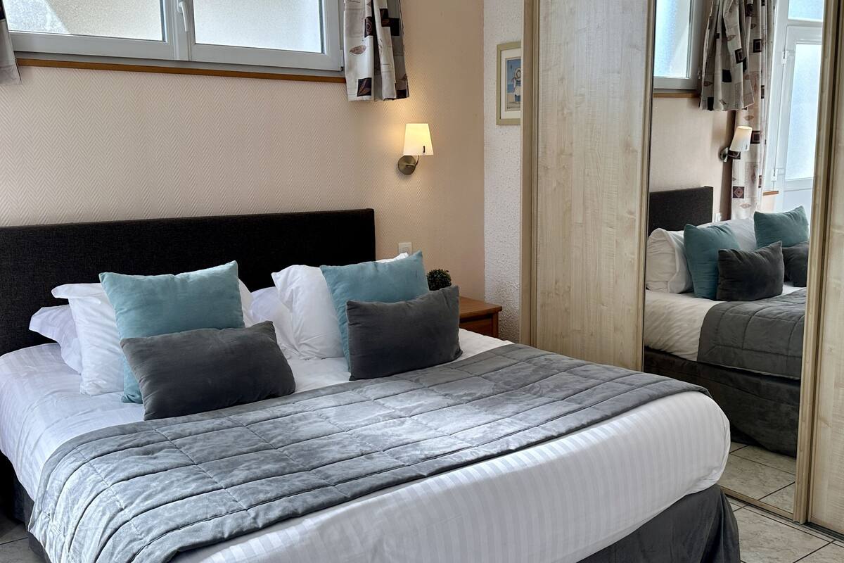A neatly made bed with a grey quilt and multiple pillows, a bedside lamp, and a mirror reflecting the room.