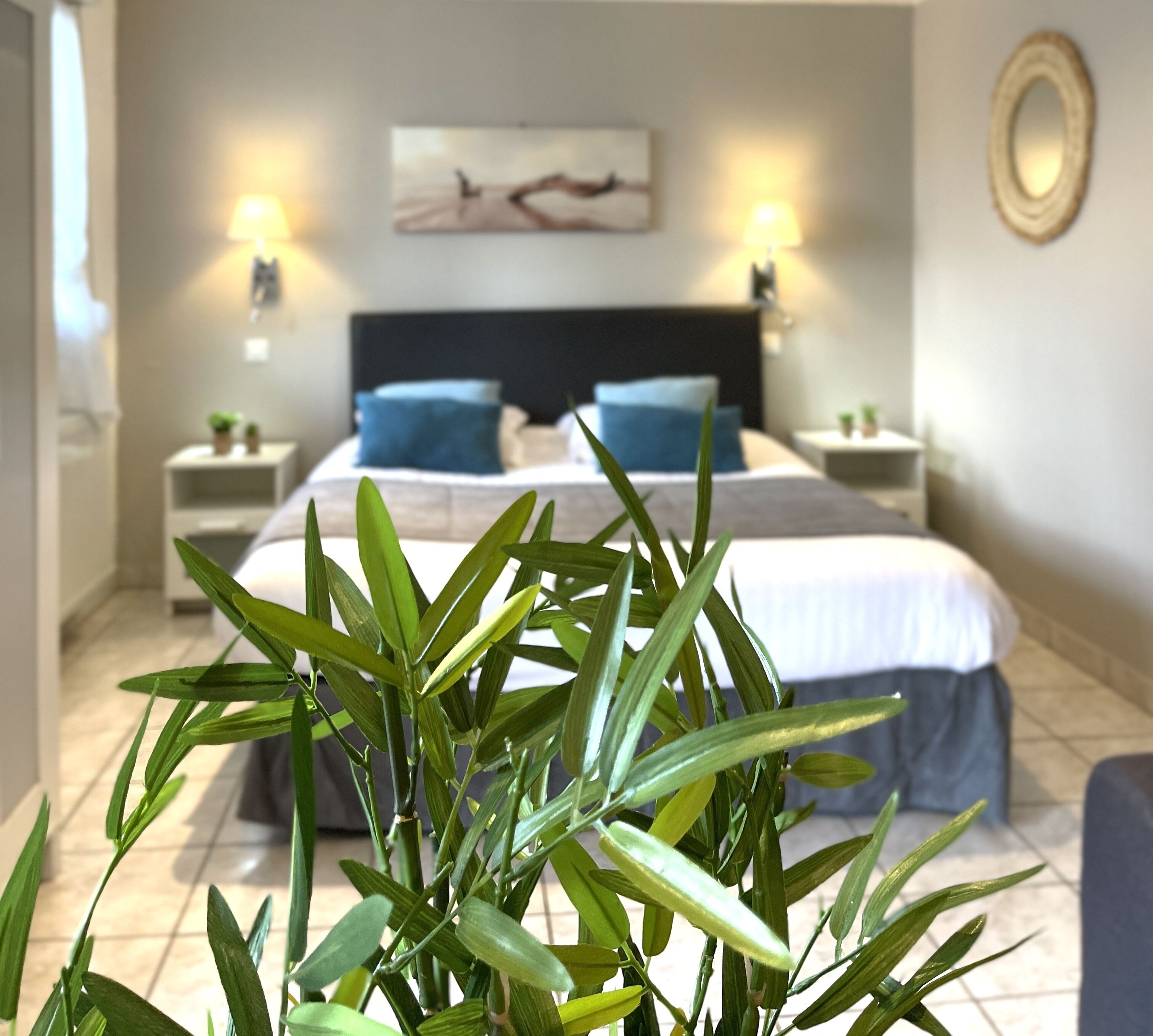 A hotel room with a bed, two bedside tables, a nightstand, a lamp, a painting on the wall, and a potted plant in the foreground.