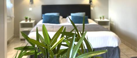 A hotel room with a bed, two bedside tables, a nightstand, a lamp, a painting on the wall, and a potted plant in the foreground.