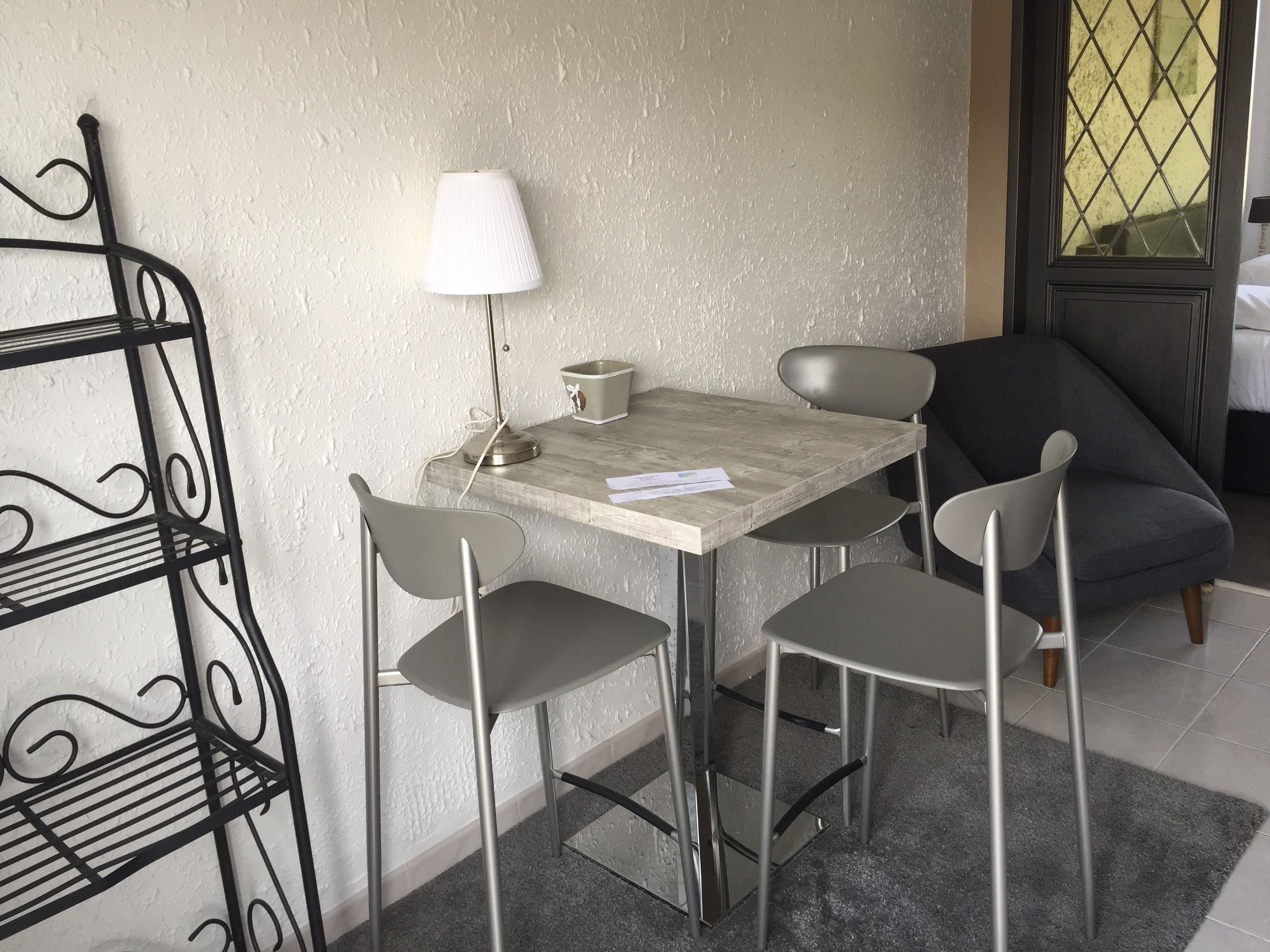 A small dining area with a table and four chairs, a metal shelf unit, a lamp, and a window with a view.