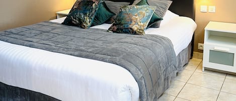 Uma cama bem arrumada com um edredom cinza e almofadas, uma mesinha de cabeceira branca com uma luminária, e um armário branco.