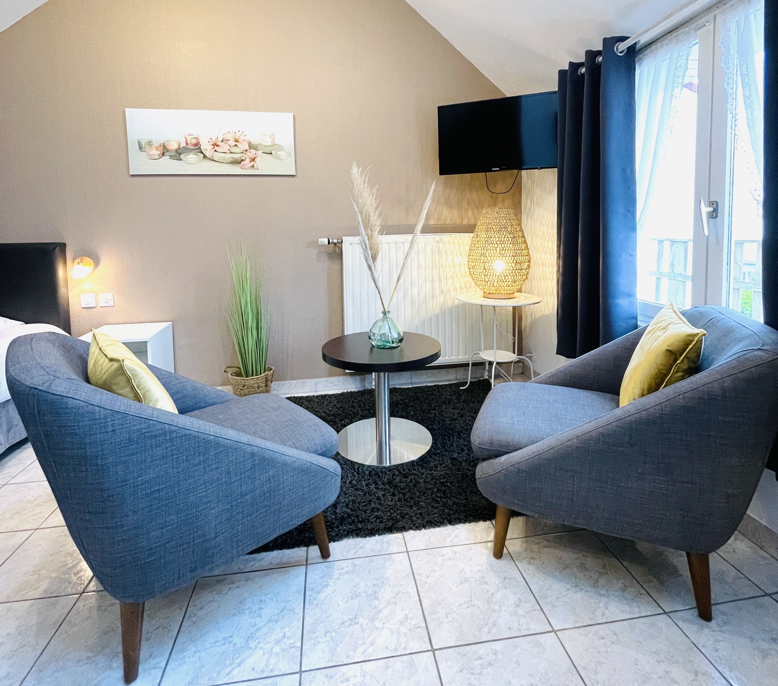 A modern living room with two grey armchairs, a round table with a vase, a flat-screen TV mounted on the wall, and a large window with curtains.