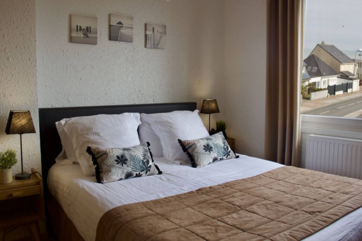A neatly made bed with white linens and two decorative pillows, a bedside table with a lamp, and a view of a street through a window.
