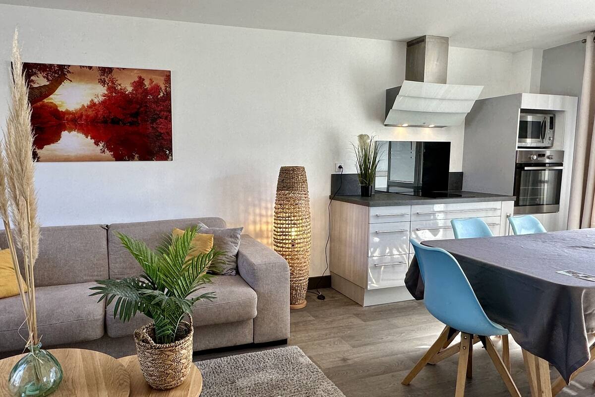 A modern living room with a grey sofa, a dining table with blue chairs, a kitchen with a microwave and oven, and a tall woven lamp.