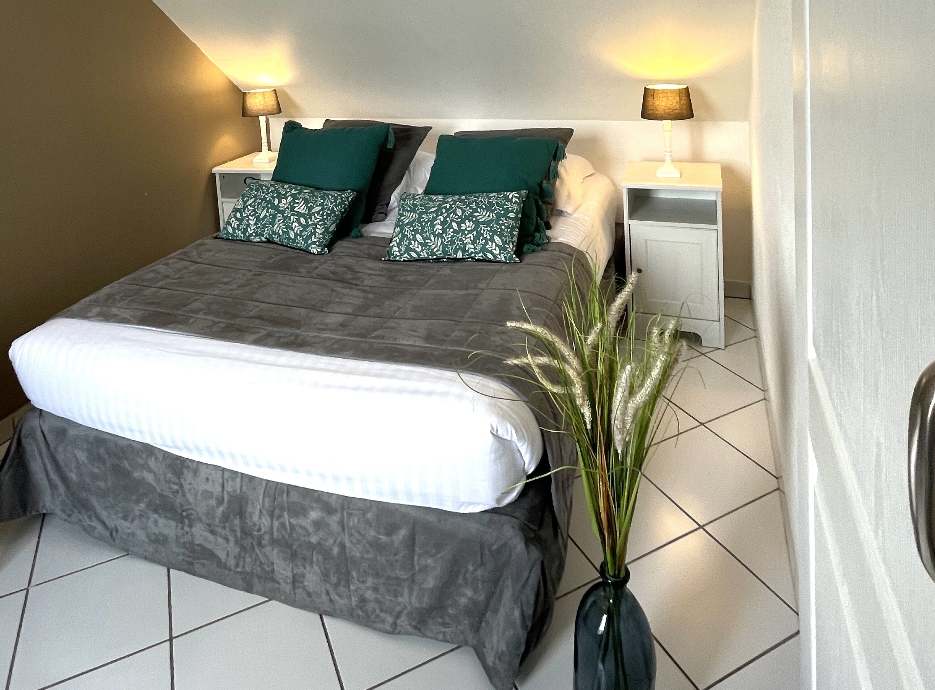 Un lit bien fait, avec une couette grise et blanche, deux oreillers verts et une lampe de chevet de chaque côté.