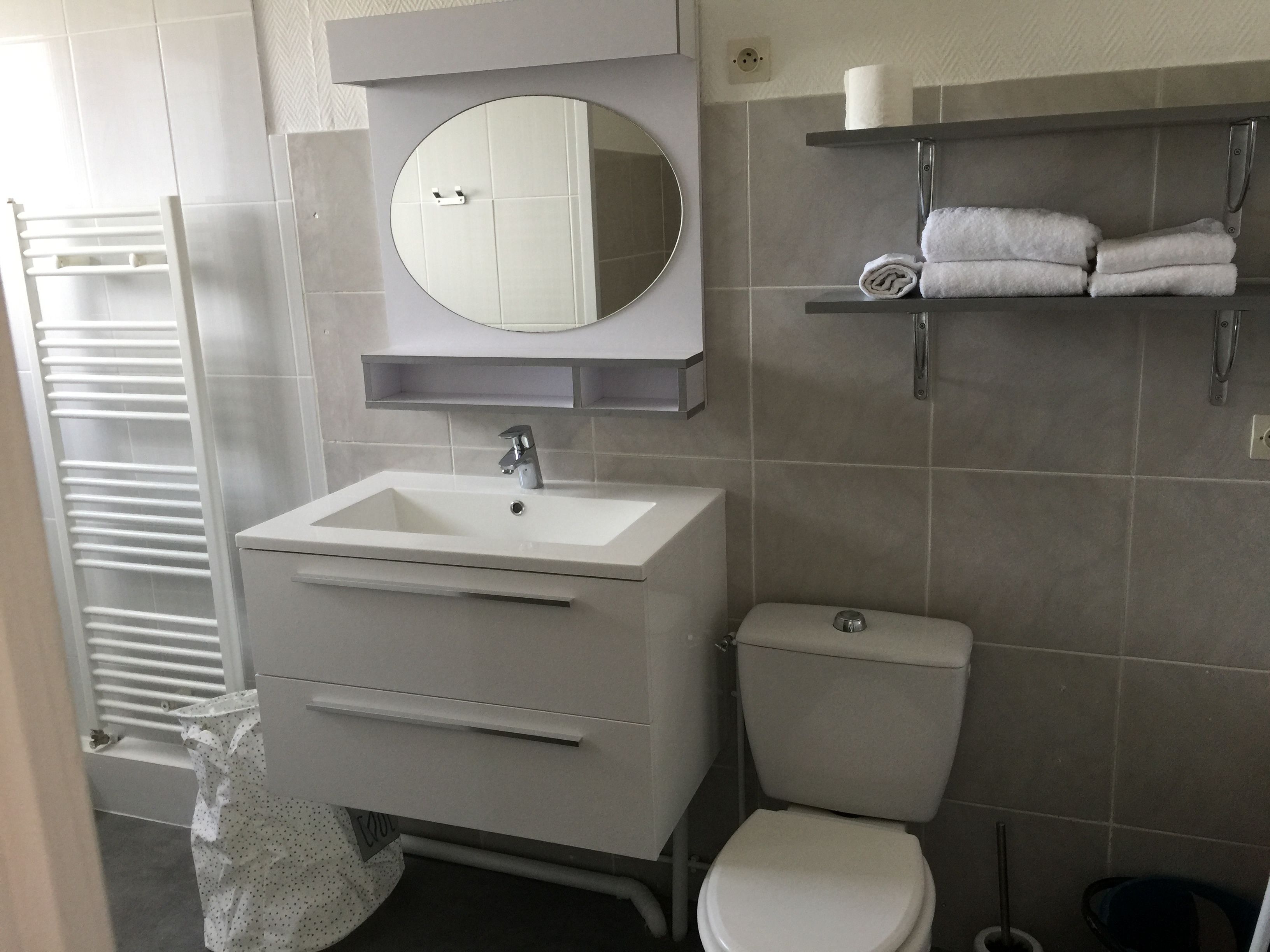 Une salle de bain moderne avec un lavabo blanc, un miroir et des toilettes. On trouve un porte-serviettes au-dessus des toilettes et une étagère avec des serviettes.