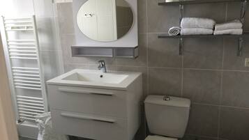 Une salle de bain moderne avec un lavabo blanc, un miroir et des toilettes. On trouve un porte-serviettes au-dessus des toilettes et une étagère avec des serviettes.