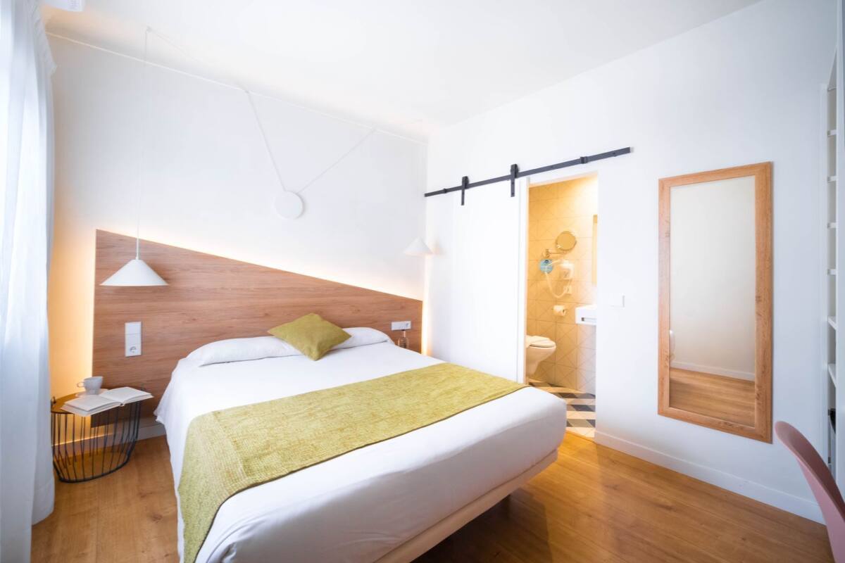 Habitación de hotel con cama, mesita de noche, lámpara y baño a la vista a través de una puerta abierta.