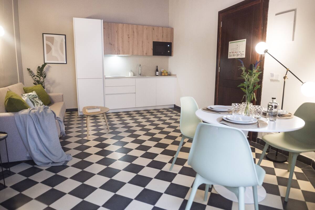 Un espacio habitable compacto con kitchenette, un comedor con mesa y sillas redondas, y un sofá con cojines.
