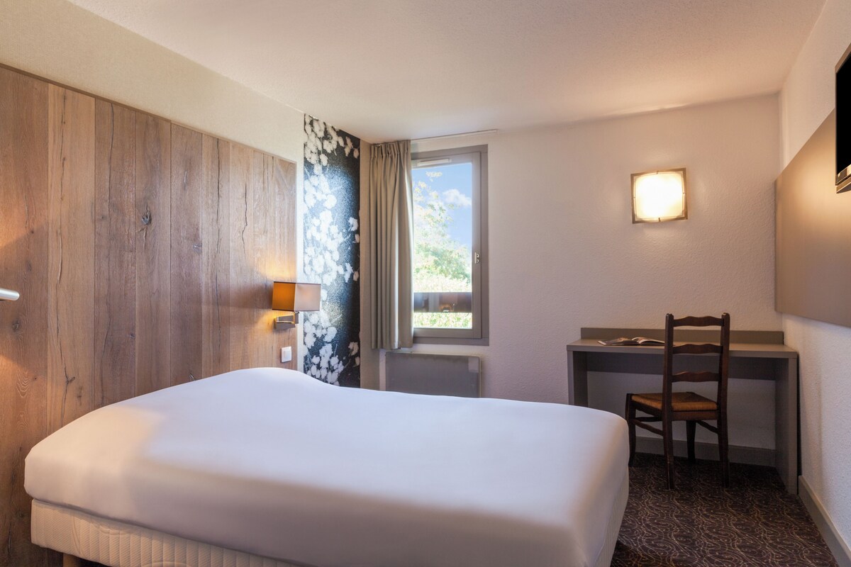 A hotel room with a bed, a desk, a chair, a window with curtains, and a wall-mounted light.