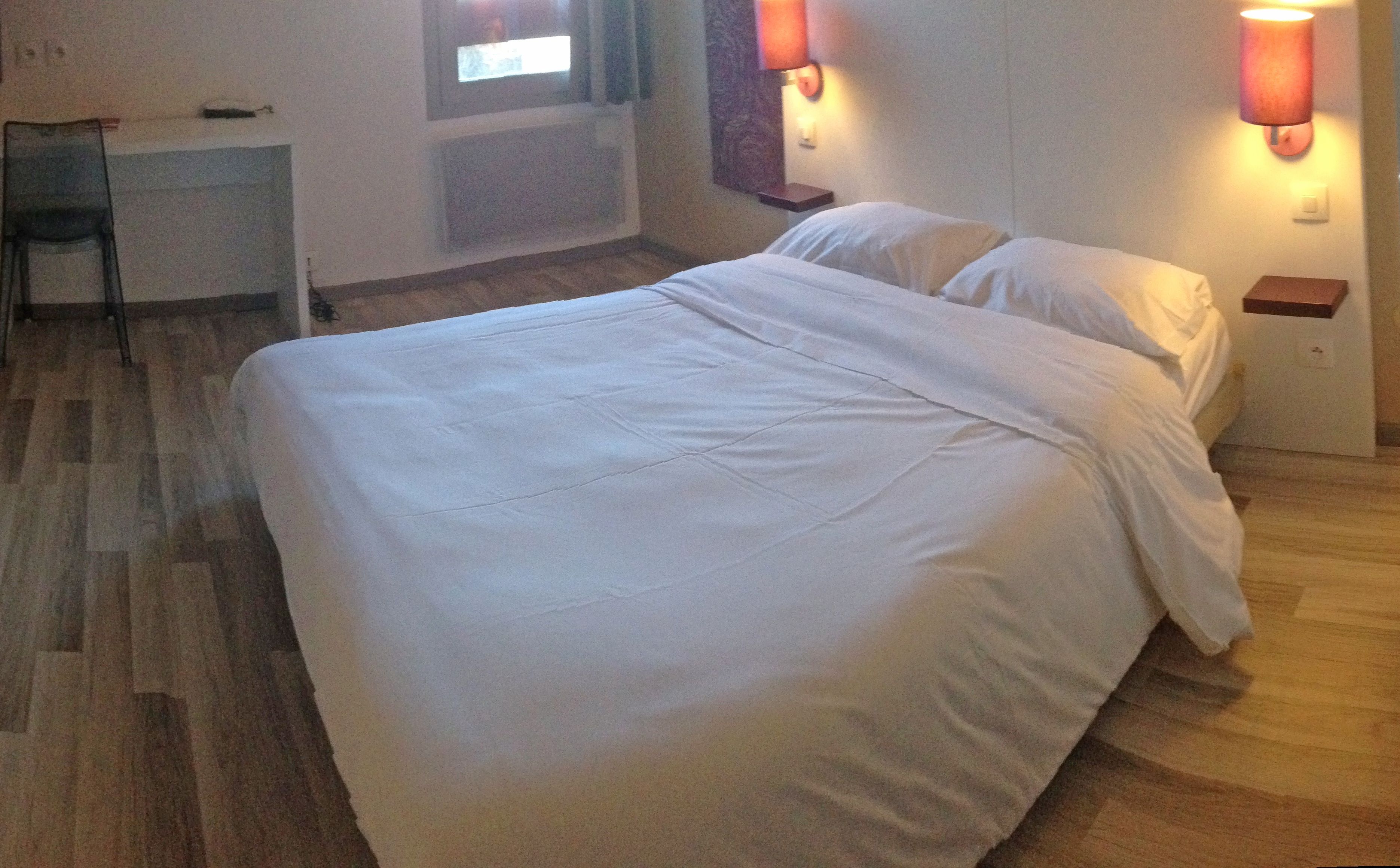 A hotel room with a large bed, a desk, and two wall-mounted lamps.