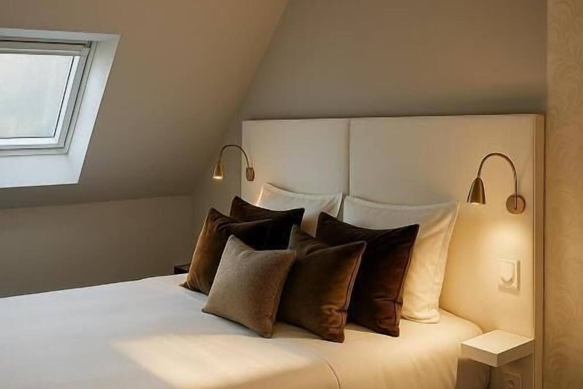 A bedroom with a bed, pillows, a nightstand, a wall-mounted lamp, and a window.