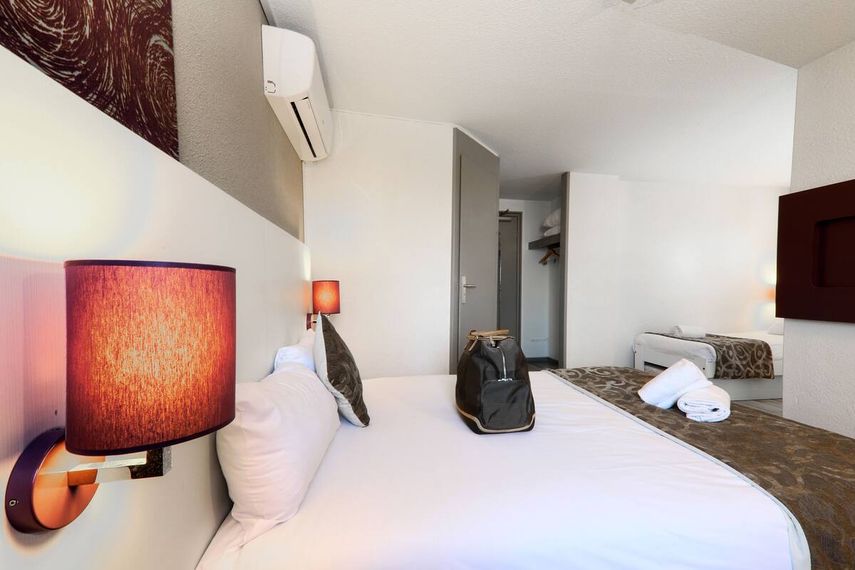 A hotel room with a bed, a wall-mounted lamp, a suitcase, and a door leading to another room.