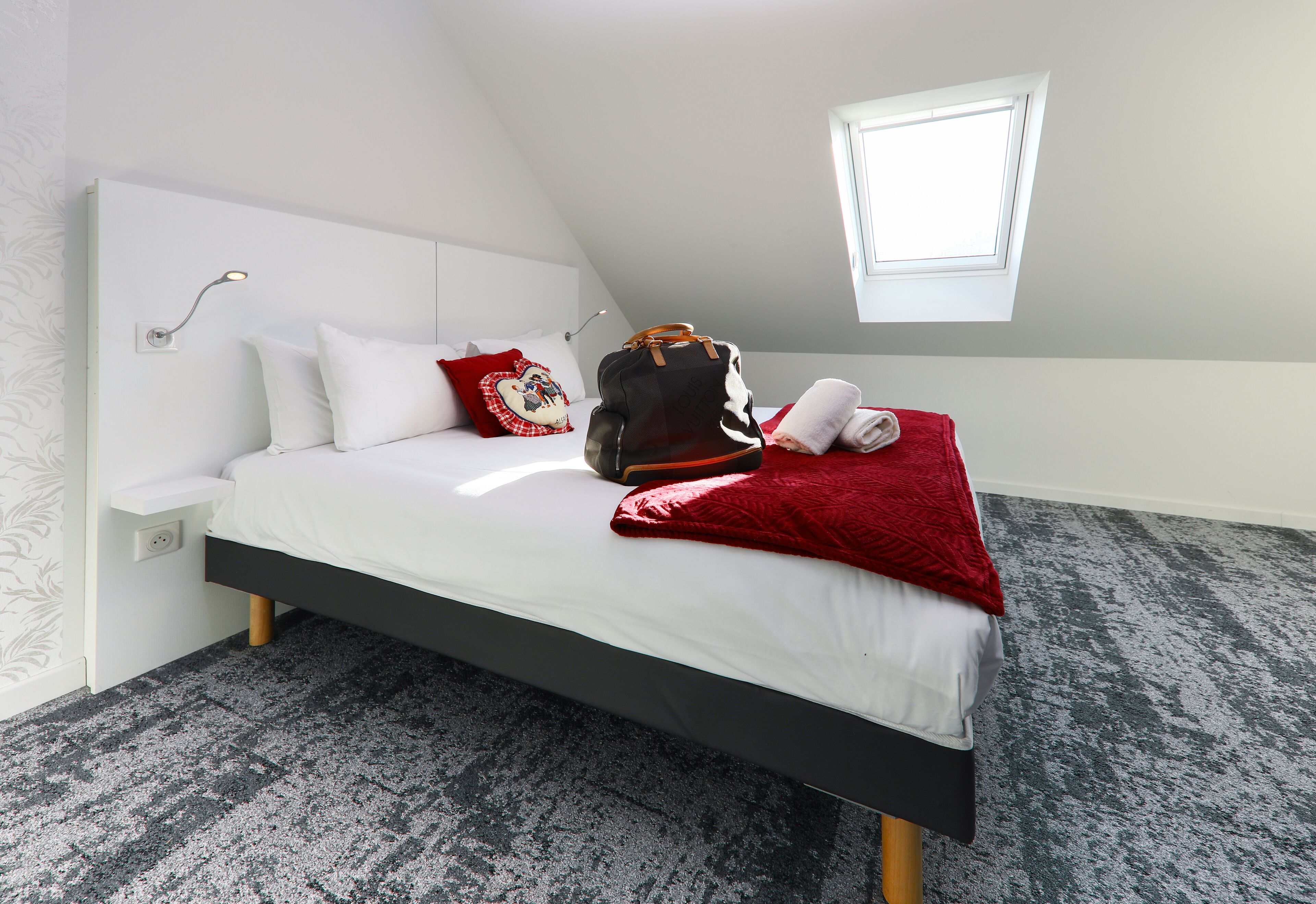 Un dormitorio moderno con una cama, una manta roja, una mochila y una ventana pequeña.