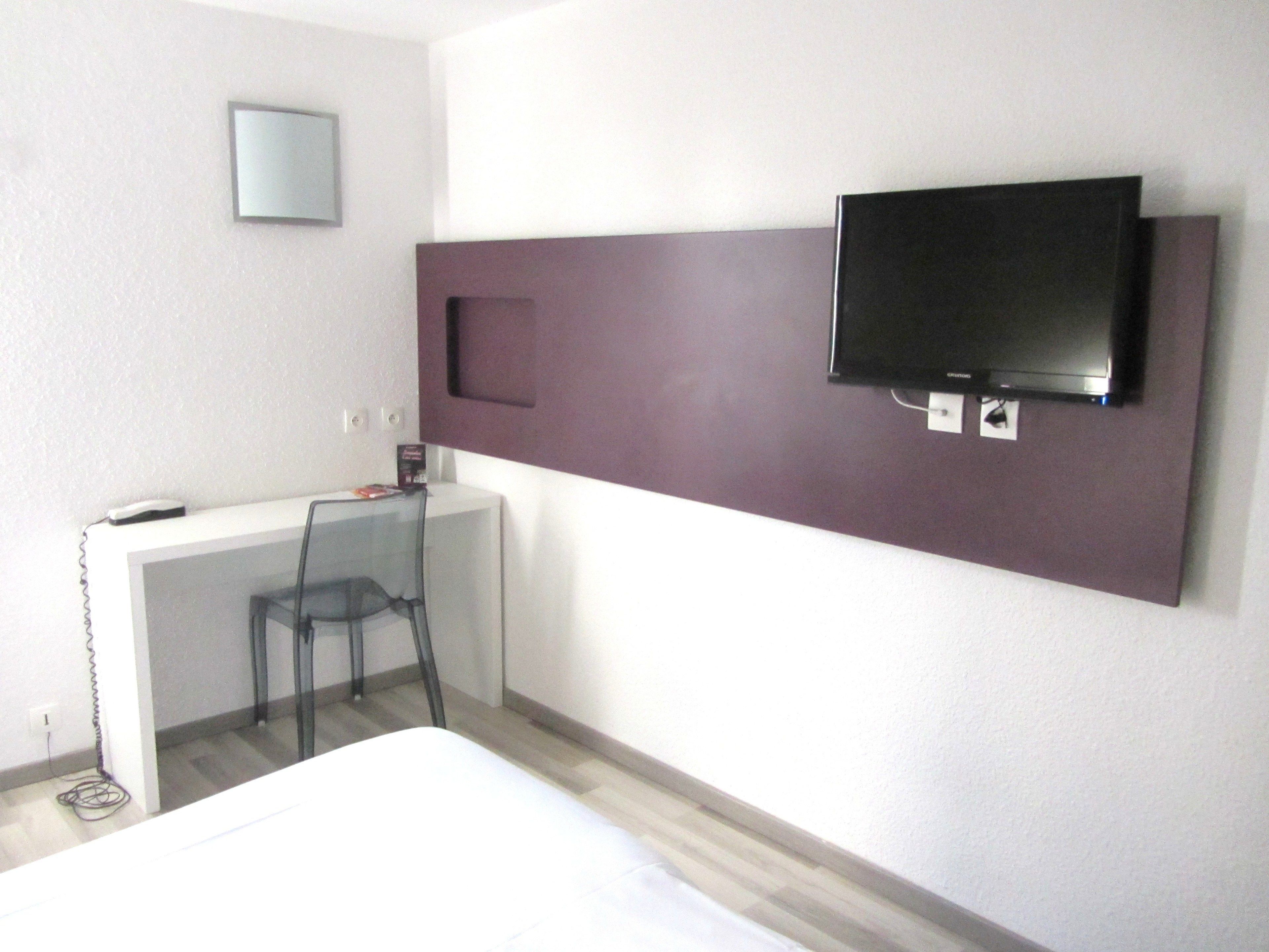 Una habitación de hotel con una cama, un escritorio con una silla, un televisor montado en la pared y un espejo pequeño sobre el escritorio.