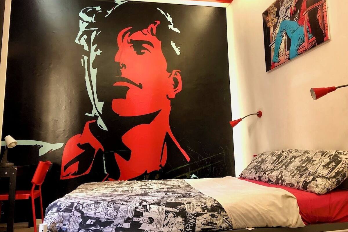 A bedroom with a large mural of a person, a bed with a patterned blanket, a red chair, and a red lamp.
