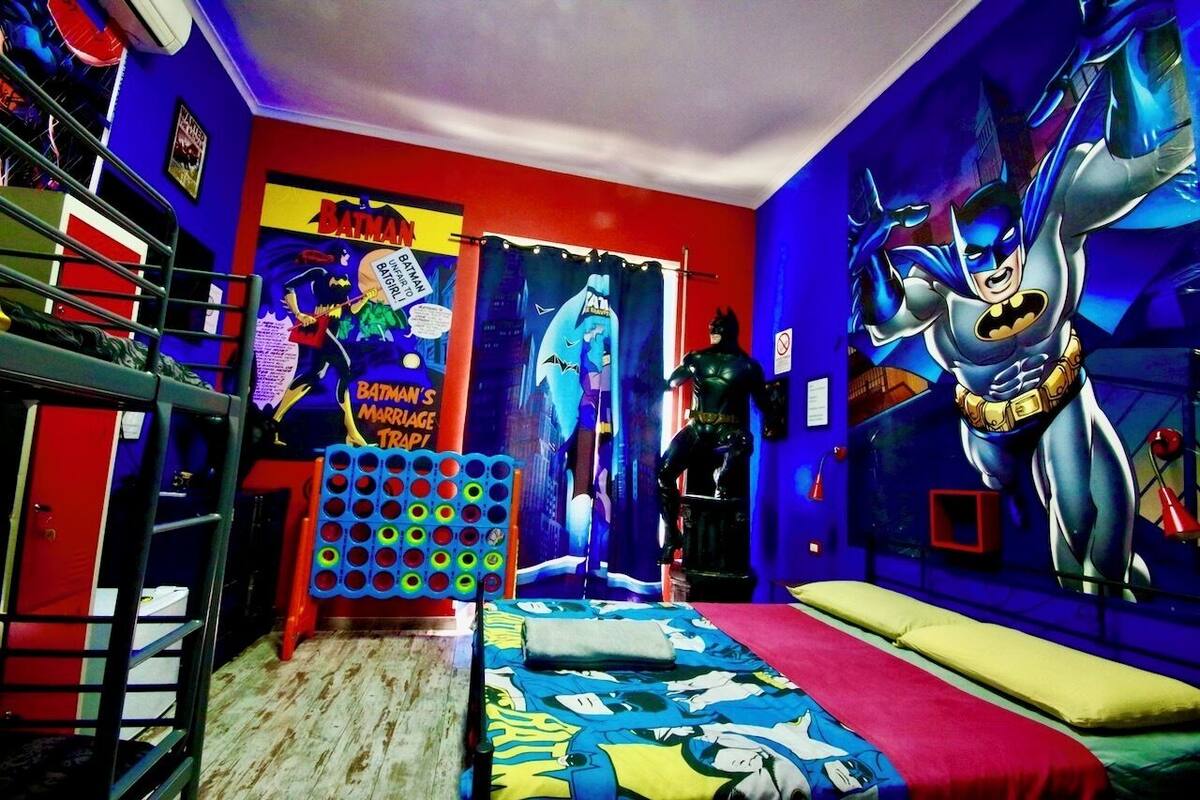 A room with a Batman-themed mural, a bed with a colorful Batman-themed bedspread, a small table with a Batman-themed toy, and a Batman-themed wall decoration.
