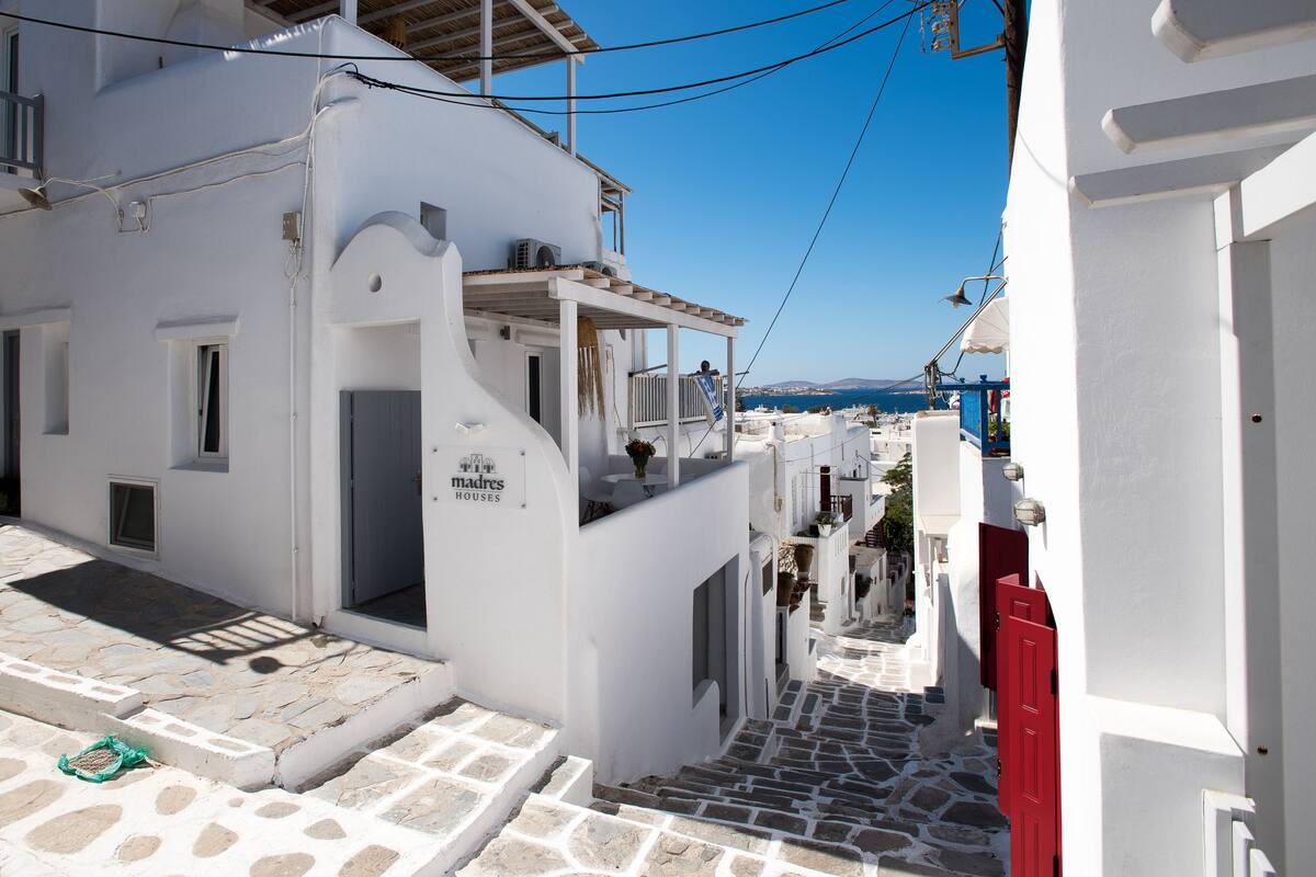 A narrow street with white buildings, a visible sign for 'Madres Houses', and a clear view of the sea in the background.