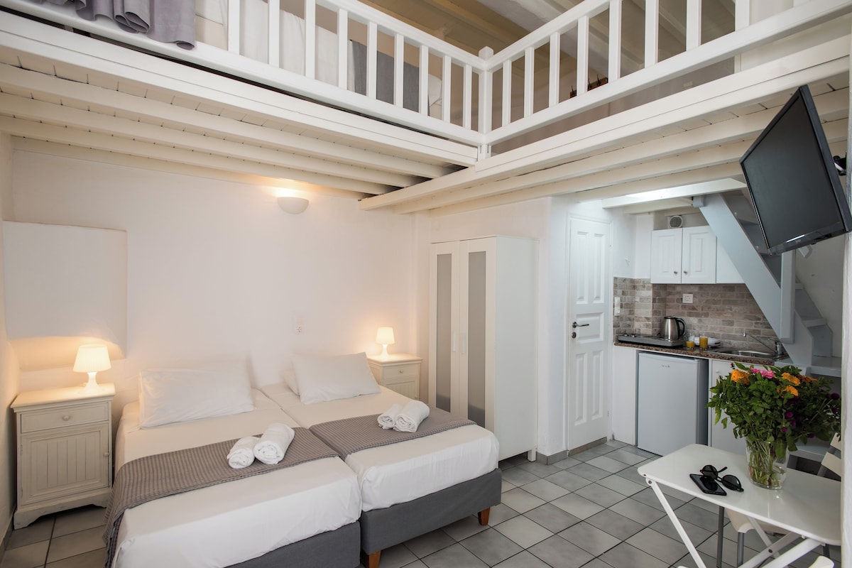 A bedroom with two beds, a small table with a vase of flowers, a kitchenette, and a balcony.